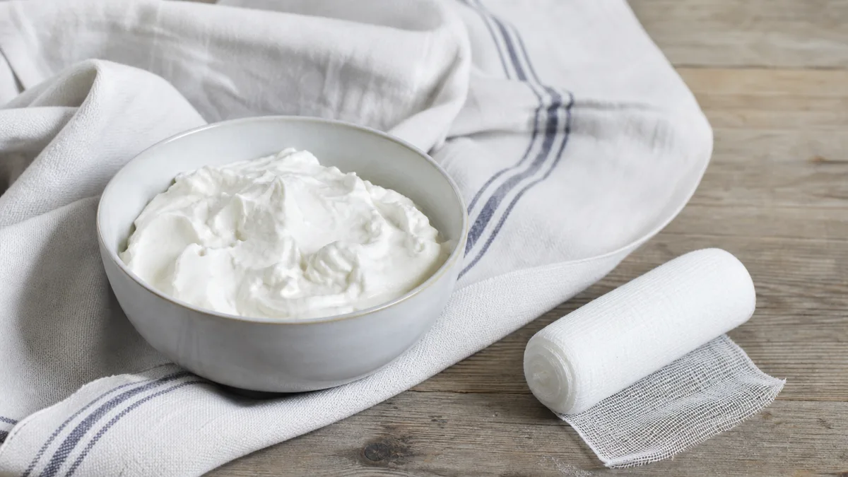 Frischer Quark auf einem dünnen Baumwolltuch neben einem Knie auf heller Unterlage, daneben ein Frottiertuch und ein Löffel